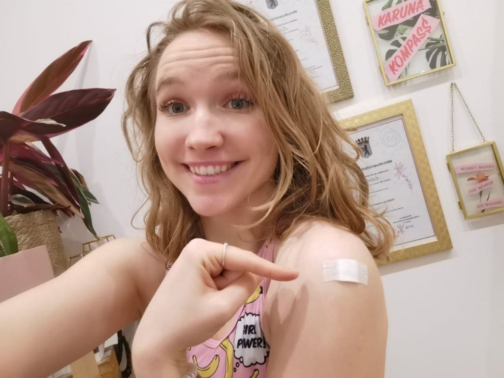 Luise Lehmann im Selfie nach der Impfung, ein Pflaster klebt auf dem Oberarm, sie zeigt mit dem Finger darauf und lächelt breit.